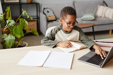 Portrait of smiling African-American boy wearing glasses using laptop while studying at home,...