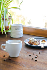 white espresso coffee cup, fresh baked eclair sweet dessert on plate kitchen table against window, utensils dishware, home green plant. morning french home breakfast