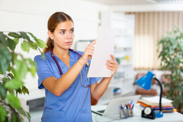 Young woman doctor working in the clinic is attentive studying the patient's outpatient card, standing in the resident's ..office. Close-up portrait..