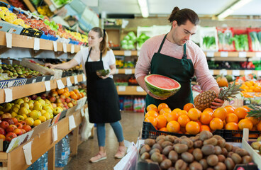 Young salesman working with fresh fruits in fruit store, woman on background