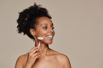 Minimal portrait of young African-American woman using face massager while enjoying skincare against neutral background, copy space