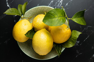 Zenital shot of a fruit bowl full of fresh and organic lemons without chemical treatment. The bowl is placed on a black marble surface