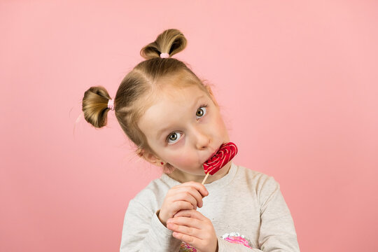 Funny little blonde girl smiling and licking red heart shape lollipop caramel on pink background. Valentines Day or Mothers Day concept