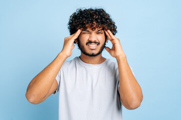 Upset unhealthy indian guy, grimacing in pain in his head, massaging his temples, experiencing a migraine, headache, overworked, in need of rest or treatment, stands on an isolated blue background