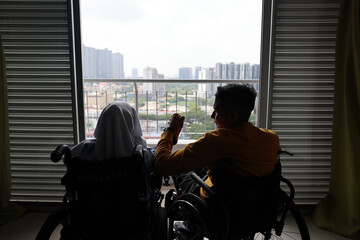 South east Asian malay man woman couple headscarf tudung middle aged disabled on wheelchair looking sitting in front of balcony window look outside holding hand