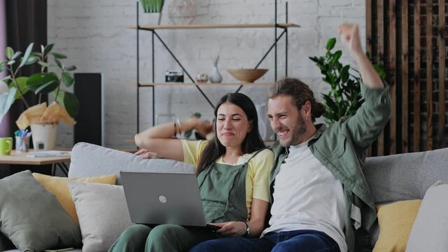 Excited young family couple winners sport fans watching game on laptop celebrating victory goal sit on couch. Overjoyed boy and girl shoppers winning online shopping voucher feeling amazed together.