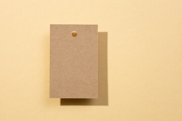 Fototapeta premium One blank cardboard label tag for clothes with tiny hole in upper part put a bit left from center with shadows falling on flesh colored background. Tag mock up. Copy space.