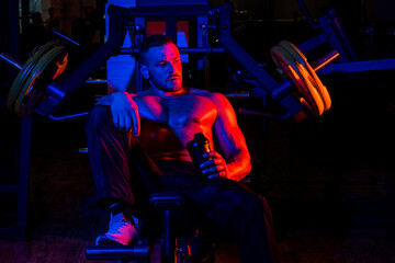 Fototapeta na wymiar Tired sporty man resting after training. Man with bottle of water or protein on neon background. Shirtless man man with muscles torso, in the gym.