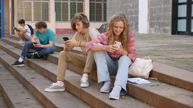 Group of teenage friends holding phones and browsing sitting on the stairs near school