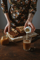 A woman in a floral print dress with a tattoo pouring milk from a white jug into a glass of black coffee. Chocolate banana bread on a wooden cutting board. A jar of salted caramel. Wooden table.