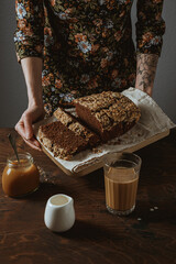 A woman in a floral print dress with a tattoo holding a wooden cutting board with chocolate banana bread. A glass of coffee with cream, a milk jug and a jar of salted caramel. Wooden table.