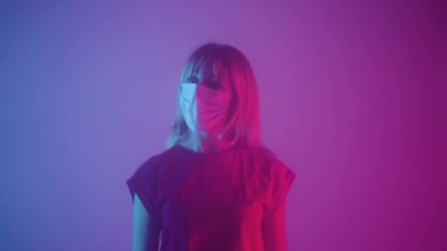 Woman in medical mask emerges from smoke and touches glass with palms. Blonde slides hands over it with upset and afraid. Model poses in dark studio with pink and purple lights in background. Close up