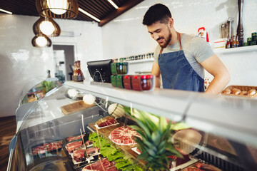 Young man butcher arranging meat products in display case of butcher shop