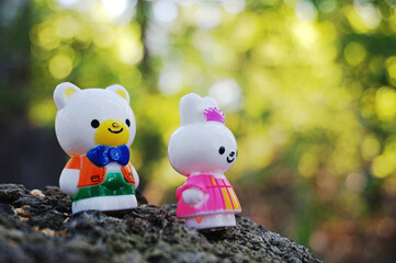 Miniature figures of a bunny and a teddy bear are standing in the forest on the ground. Shooting from below. A blurry image of trees in the background. Close-up.