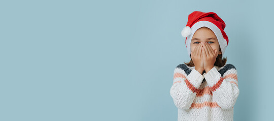 Laughing little girl in christmas hat covering her face over blue