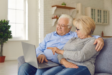 Happy senior couple shopping online, reading news and surfing internet using laptop while sitting on sofa at home. Technology connect and modern elderly lifestyles concept.