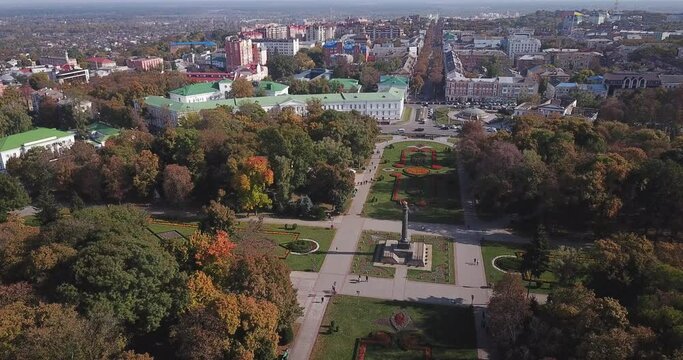 A drone descends in the central park of the Ukrainian provincial city of Poltava. Day, sunny. Green trees in the foreground, historical buildings in the background.