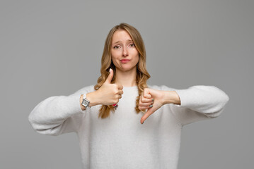 Fototapeta premium Puzzled blonde girl showing thumb up and down. Skeptical thinking woman judging deciding how to rate a situation, wears white sweater, gray background