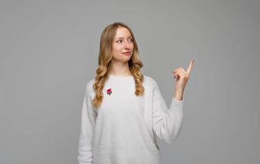 Confident successful blonde young woman finger at upper right corner and showing advertisement, standing in white sweater against gray background