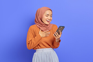 Portrait of smiling beautiful Asian woman in brown sweater and hijab holding on mobile phone with hand on chest isolated over purple background. People islam religious concept