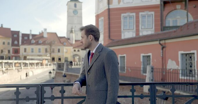 handsome businessman holding hand on bridge, looking to side, admiring city, touching face and smiling when camera is zooming in 