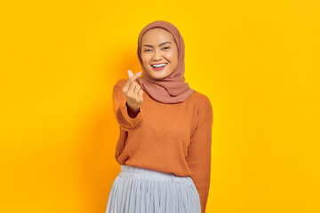 Beautiful smiling Asian woman in brown sweater showing korean heart with two fingers crossed, express joy and positivity isolated over yellow background