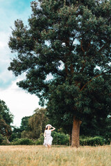 Hppy young woman in a dress and a straw hat runs across the field. In the background is a large tree and a stormy sky. Vertical