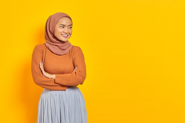 Beautiful smiling Asian woman in brown sweater and hijab crossed arms chest and looking at copy space isolated over yellow background. People islam religious concept