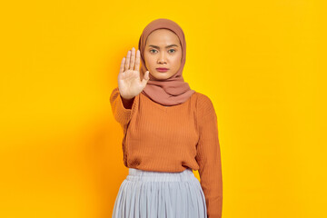 Beautiful serious Asian woman in brown sweater and hijab standing with outstretched hand showing...