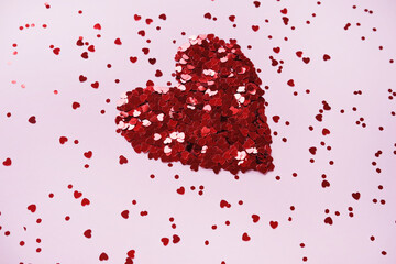 Heart made of shiny red small decorative hearts on a pink background strewn with sparkles. Side view, space for text. Valentine's Day card.
