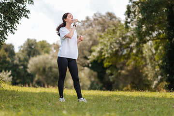 Wellness. A young sporty woman in sportswear drinking bottle of water. In the background, a summer green park