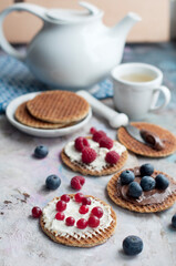 Waffles with cream cheese and chocolate, cranberries, raspberries and blueberries on a gray background. Romantic breakfast with teapot, cup of tea, knife, fruits on waffles