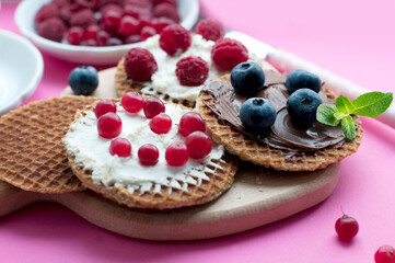 Waffles with cream cheese, cranberries, raspberries, blueberries, chocolate and mint on a wooden board on a pink background. Romantic breakfast with fruits on waffles and pink knife