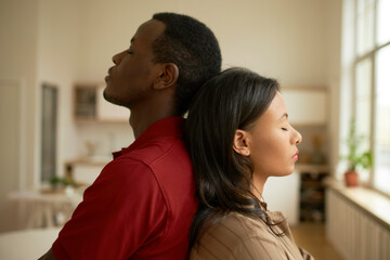 Obraz premium Picture of black man and woman standing back to back with eyes closed, feeling exhausted and offended after sorting things out isolated on blurred background of living-room. Human relationships