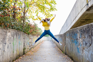 Carefree young woman in yellow jumper jumping at concrete pedestrian walkway in autumn park