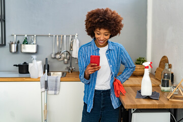 Smiling young Afro woman text messaging on smart phone by spray bottle at kitchen counter