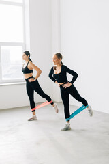 Fototapeta na wymiar Two smiling athletic Women are enjoying leg and buttocks work out with resistance band training together with friend in gym concept
