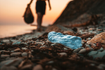 Close up of dirty blue plastic bottle lying on the beach. Defocused silhouette of a person holding...