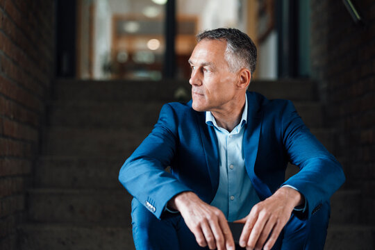 Thoughtful mature businessman sitting on steps at entrance of office building - Powered by Adobe
