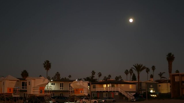 Palm trees silhouettes and full moon in twilight sky, California beach, USA. Beachfront houses or homes on coast in evening, fullmoon on pacific ocean shore in dusk. Lifeguard tower. Light in windows.
