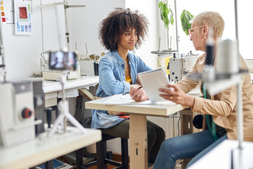 Positive African-American lady listens to colleague with sketchbook in light fashion studio