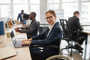 Vertical side view portrait of young businesswoman using wheelchair while working with laptop in office