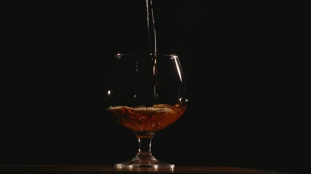  a cognac glass stands on a table against a black uniform background and cognac is poured into it
