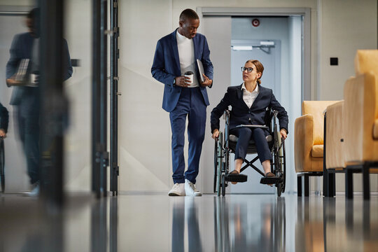 Full length portrait of successful businesswoman using wheelchair while moving towards camera and talking to colleague in modern office space