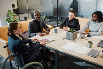 High angle portrait of successful businesswoman using wheelchair at meeting and talking to colleagues in modern office space