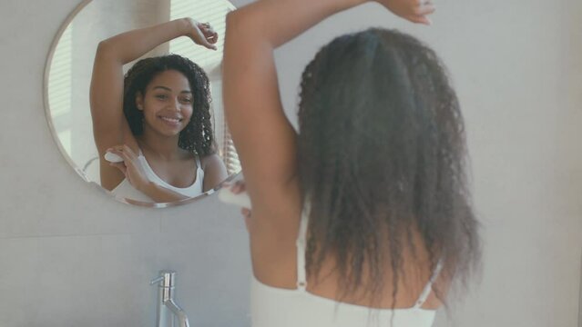 Hygienic cosmetics. Young african american woman applying stick deodorant on her armpit, looking at mirror at bathroom