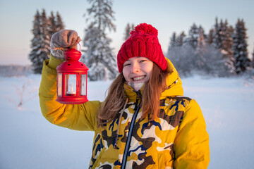 A teenage girl joyfully holds in her hands a red candlestick lantern in the winter forest.