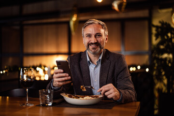 Portrait of caucasian adult man, smiling for the camera, while holding his phone and eating.