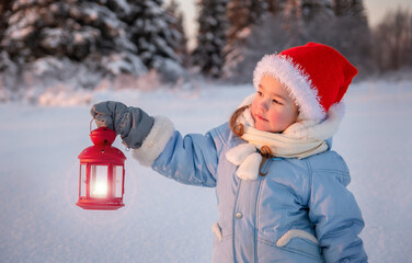 In winter, in the forest, a girl in a blue jacket and a red cap holds a red lantern in her hands. The concept of a Christmas child.