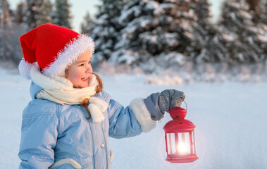 In winter, in the forest, a girl in a blue jacket and a red cap holds a red lantern in her hands. The concept of a Christmas child.
